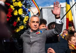 ălin Georgescu salutând mulțimea la un eveniment public, îmbrăcat într-o jachetă gri cu insigna tricoloră, înconjurat de flori și steaguri românești, simbolizând sprijinul popular în campania sa