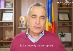 Călin Gergescu stă în fața unei cămări cu cărți, vorbind în timpul unui interviu video. În fundal se observă un steag al României și diverse obiecte decorative.