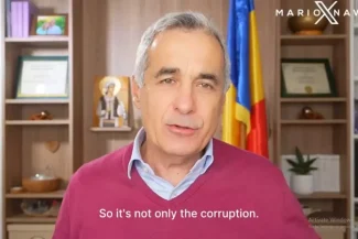 Călin Gergescu stă în fața unei cămări cu cărți, vorbind în timpul unui interviu video. În fundal se observă un steag al României și diverse obiecte decorative.