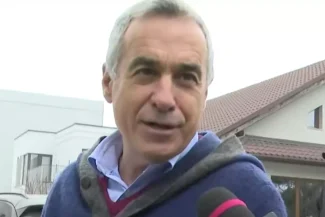 Călin Georgescu vorbind cu reporterii în exterior, purtând un gilet gri și o cămașă albastră, cu o expresie preocupată pe față