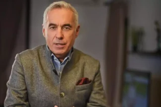 Călin Georgescu vorbește serios într-un interviu, îmbrăcat într-un sacou gri de lână, cu o eșarfă burgundy în buzunar, într-un decor interior