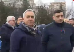 Călin Georgescu însoțit de George Simion la un eveniment public, purtând un fular și o geacă albastră, cu o expresie serioasă, discutând cu forțele de ordine la un miting.