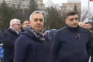 Călin Georgescu însoțit de George Simion la un eveniment public, purtând un fular și o geacă albastră, cu o expresie serioasă, discutând cu forțele de ordine la un miting.