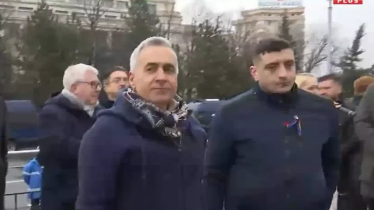Călin Georgescu însoțit de George Simion la un eveniment public, purtând un fular și o geacă albastră, cu o expresie serioasă, discutând cu forțele de ordine la un miting.