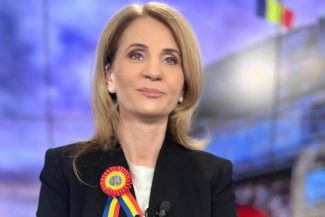Anca Alexandrescu, prezentatoarea emisiunii Culisele Statului Paralel, zâmbind în timp ce poartă o cocardă tricoloră, reprezentând demnitatea și patriotismul în mass-media românească.