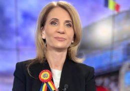 Anca Alexandrescu, jurnalista Realitatea PLUS, surâzând în studio, purtând o rosetă tricoloră.