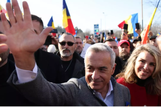 Călin Georgescu salută susținătorii în timpul unui miting, cu multitudine de steaguri românești în fundal