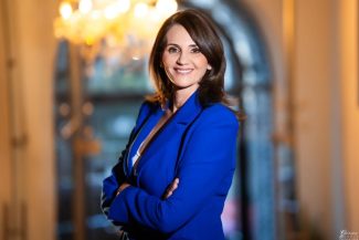 Anca Alexandrescu într-un portret oficial, purtând un sacou albastru, zâmbitoare, pe un fundal elegant