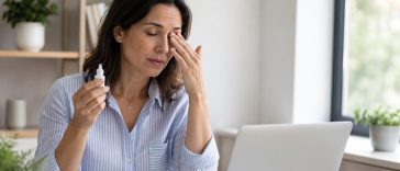 Femeie cu simptome de ochi uscat la birou, ținând picături oftalmologice lângă laptop
